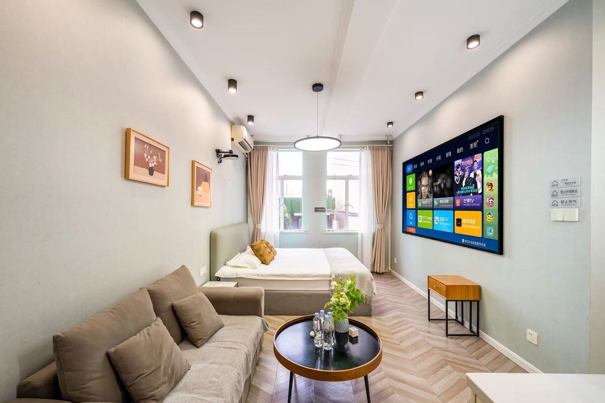 Une chambre d’hôtel moderne équipée d’un canapé, d’une petite table avec un vase, d’un lit et d’un téléviseur fixé au mur, qui diffuse différentes chaînes.
