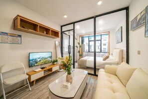 A modern hotel room with a sofa, a coffee table, a TV, a desk, and a sliding glass door leading to a balcony.