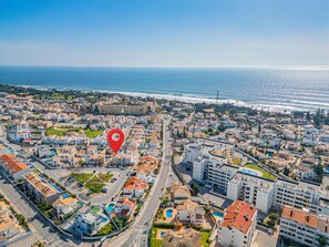 An aerial view of a coastal town with a red location marker, featuring residential buildings, a marina, and a beach.
