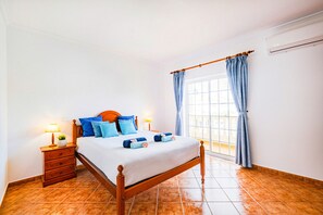 Un dormitorio con una cama de madera, una mesita de noche con lámpara, un ventanal con cortinas y suelo de baldosas.