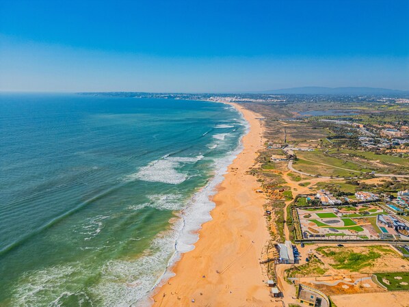 An aerial view of a coastal area with a sandy beach, a clear blue ocean, and a mix of residential and commercial buildings.