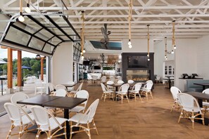A spacious restaurant with high ceilings, exposed beams, and a large glass wall overlooking the outdoor area. Tables and chairs are arranged for dining.