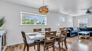 Un coin repas avec une table blanche, des chaises en bois et un luminaire suspendu. On aperçoit un canapé bleu et une petite table en bois à l’arrière-plan.