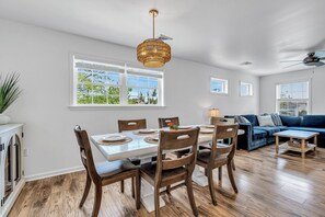 A dining area with a white table, wooden chairs, and a hanging light fixture. There is a blue sofa and a small wooden table in the background. - Luxury Bayside Ortley Beach House • Sleeps 10 (Seaside Heights)