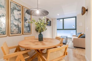 Een eetgedeelte met een ronde houten tafel, rieten stoelen, een bloemenvaas en abstracte schilderijen aan de muur.