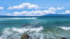 Vista costera con agua azul cristalina, olas y montañas distantes bajo un cielo parcialmente nublado.