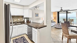 Une cuisine moderne avec des meubles blancs, un plan de travail en granit et un lave-vaisselle. On y trouve un coin repas avec des chaises en osier et une table ronde, ainsi qu’un balcon avec vue sur l’océan.