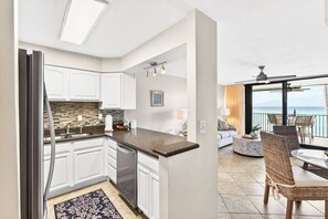 A modern kitchen with white cabinets, a granite countertop, and a dishwasher. There is a dining area with wicker chairs and a round table, and a balcony with a view of the ocean. - Coastal Elegance: A Fresh Oceanfront Gem (Lahaina)