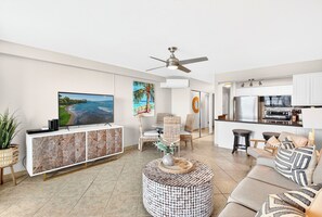 A modern living room with a flat-screen TV, a round coffee table, and a sofa. There is a kitchen area with a refrigerator and a microwave. - Coastal Elegance: A Fresh Oceanfront Gem (Lahaina)