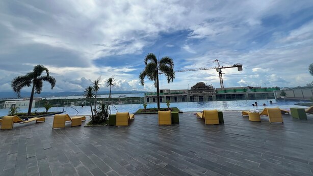 Área de piscina no terraço, com espreguiçadeiras e palmeiras, com vista para um corpo de água e um canteiro de obras.