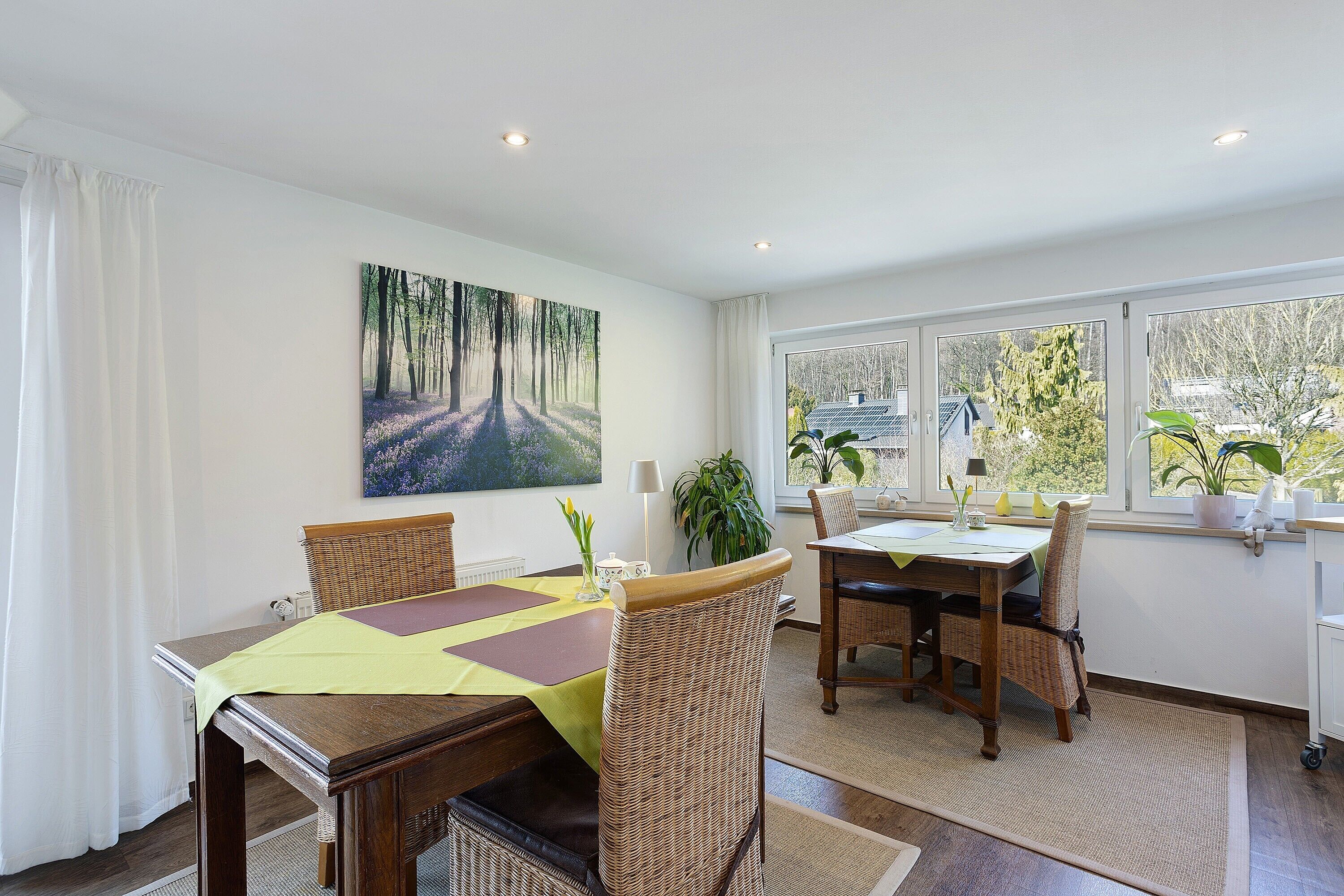 Un espace repas comprenant une table, des chaises, un tableau et une vue dégagée sur l’extérieur grâce à de grandes fenêtres.