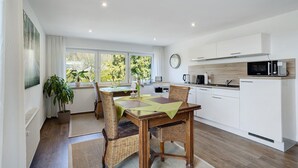 Une cuisine moderne avec des meubles blancs, une table à manger avec des chaises, une plante en pot et une vue sur les arbres par la fenêtre.