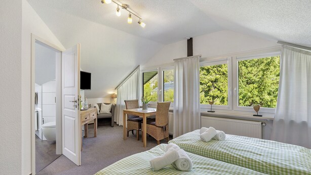 Quarto de hotel com cama, escrivaninha, cadeira e janela com vista para árvores.