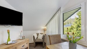 Quarto com televisão, escrivaninha com vaso e garrafa, cadeira, mesa e janela com vista.