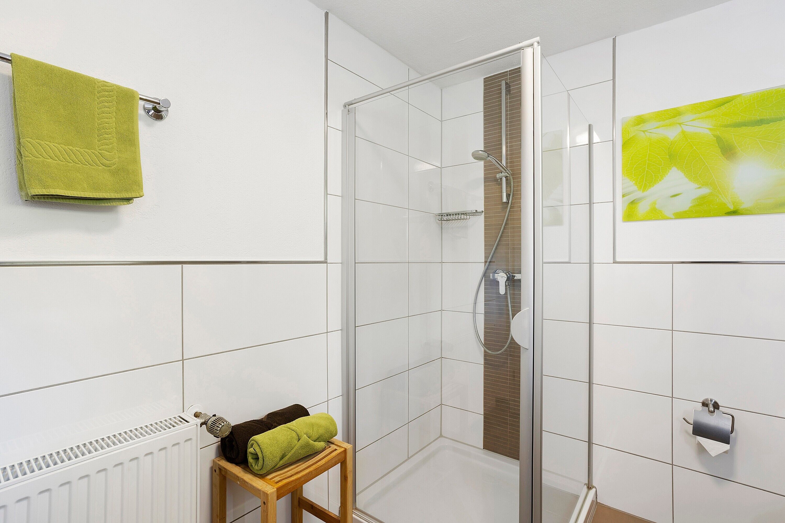 Eine Dusche mit Glastür, ein Handtuchhalter mit einem grünen Handtuch, ein hölzerner Hocker mit Handtüchern und ein Toilettenpapierhalter.