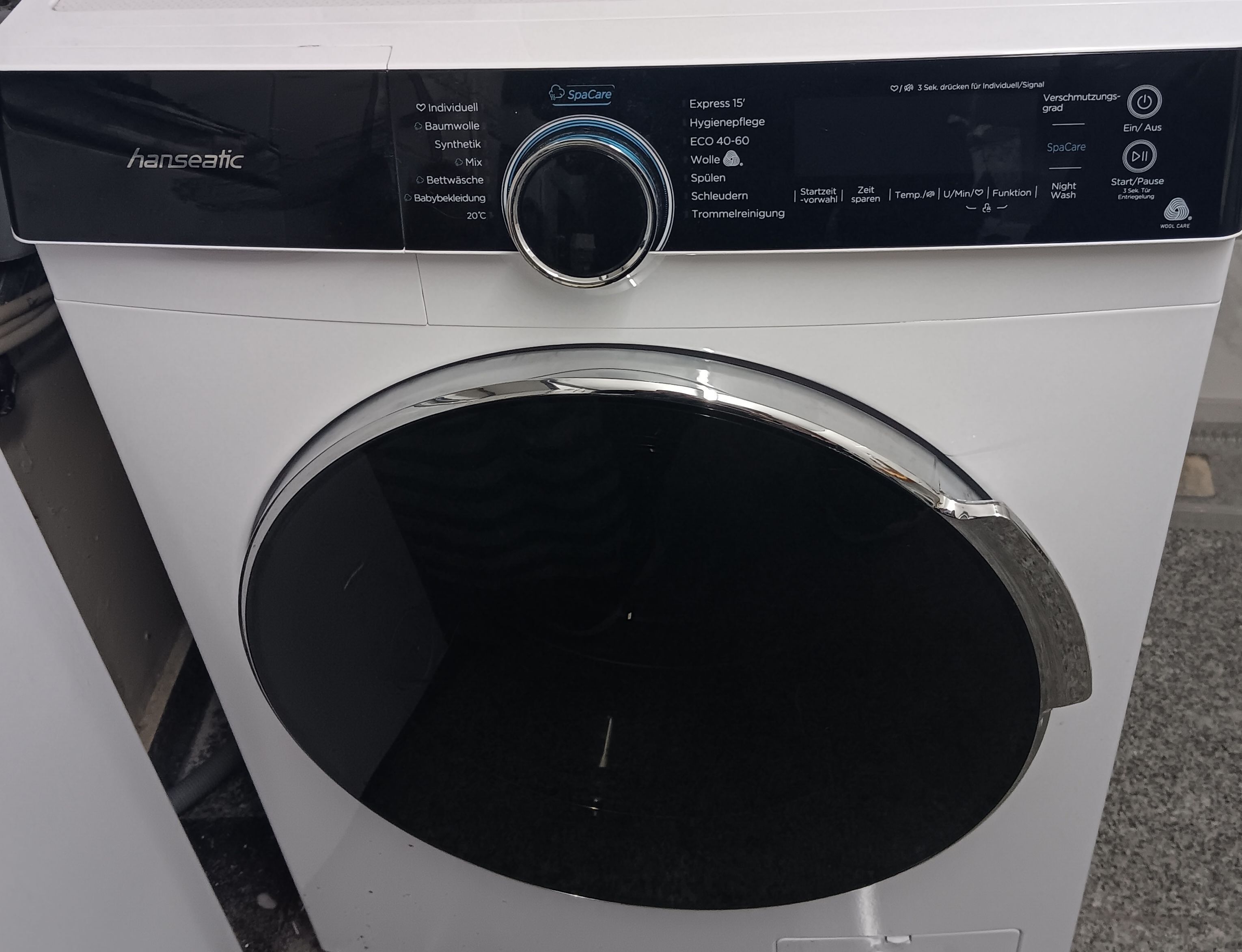 Eine Frontansicht einer weißen Waschmaschine mit schwarzem Bedienfeld, das ein Wählrad und verschiedene Tasten für unterschiedliche Waschprogramme zeigt.