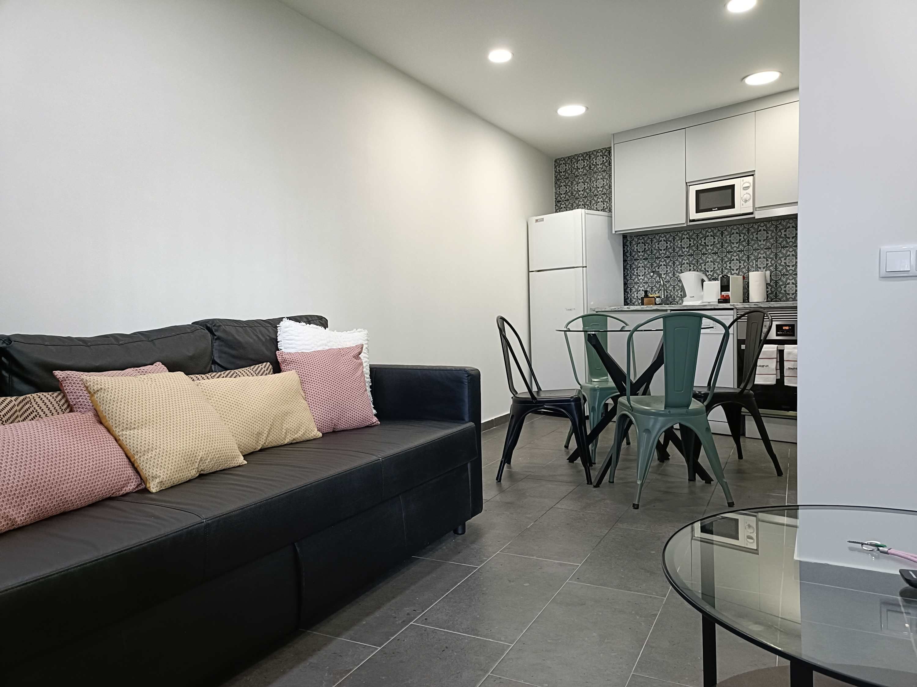 A compact living space with a black sofa, a glass dining table, and a kitchen area with white cabinets and a microwave.