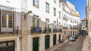 A narrow cobblestone street lined with white buildings, some with balconies and signs indicating for sale. - Super Central Walk Anywhere w AC Lift Parking (Lisbon)
