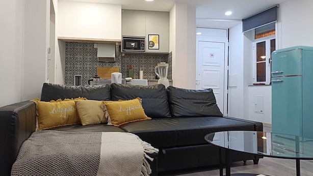 Un salón moderno con un sofá de cuero negro, una mesa de centro de vidrio y una cocina al fondo.