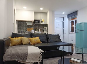 Un salón moderno con un sofá de cuero negro, una mesa de centro de vidrio y una cocina al fondo.