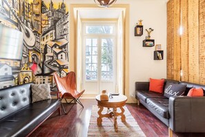 A living room with a black leather sofa, a wooden coffee table, and a red chair. There is a large window with a view of buildings outside. - Artistic Alfama 4BR Cathedral Views Tram 28 (Lisbon)