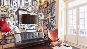 A living room with a black sofa, a round wooden coffee table, and a red chair. A large mural of city buildings is on the wall behind the sofa. - Artistic Alfama 4BR Cathedral Views Tram 28 (Lisbon)