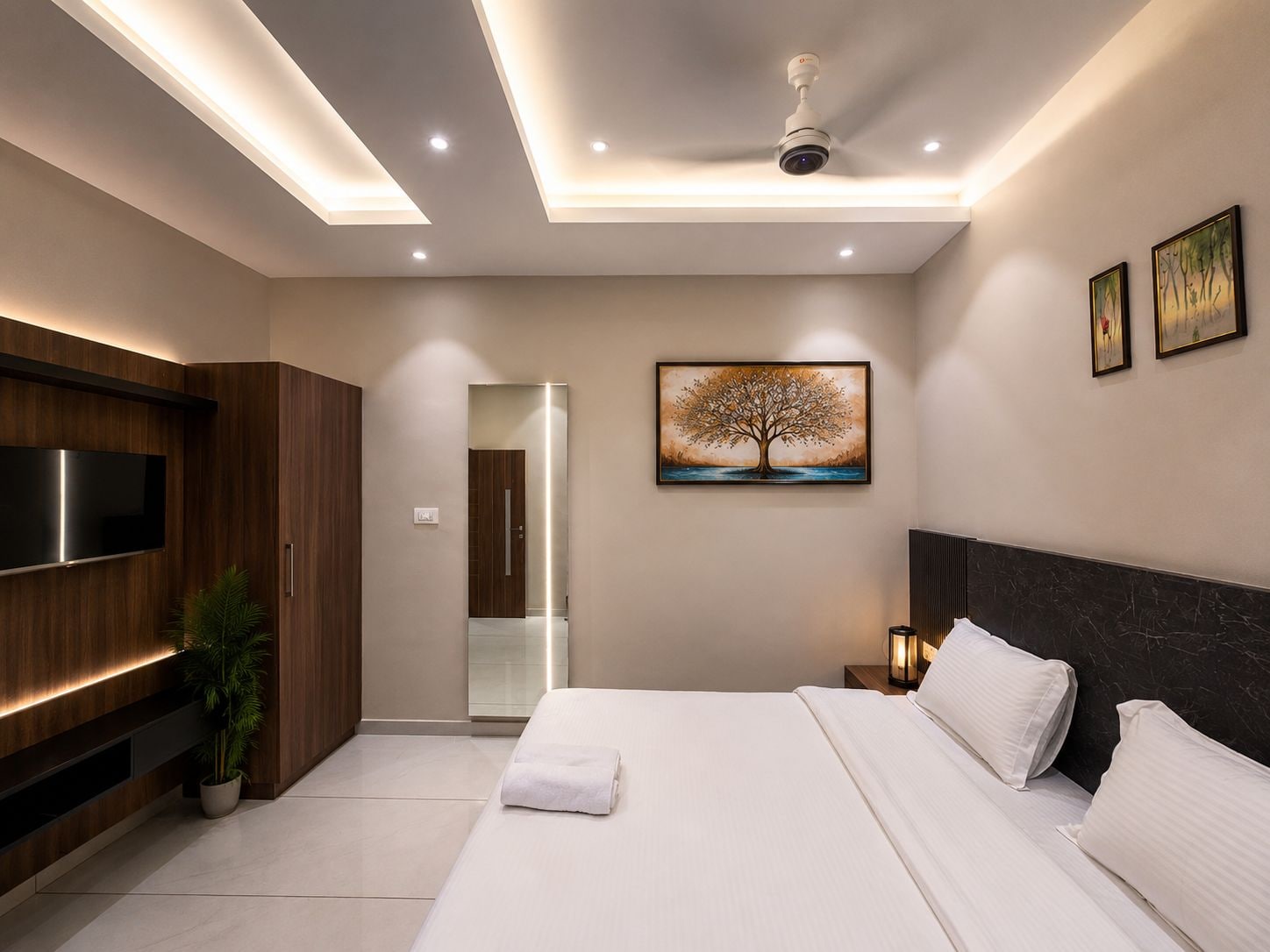 Une chambre moderne équipée d’un téléviseur à écran plat, d’un ventilateur de plafond, d’un tableau représentant un arbre et d’un lit avec du linge de lit blanc.