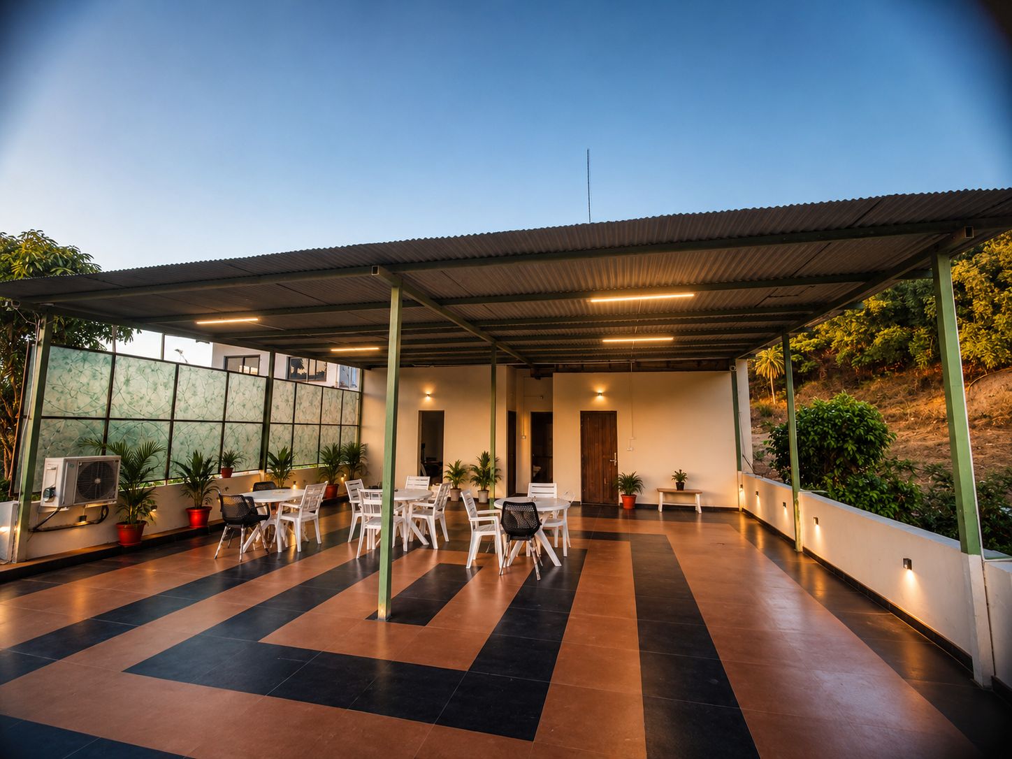 Un área de patio techada al aire libre con varias mesas y sillas, plantas en macetas y un equipo de aire acondicionado grande.