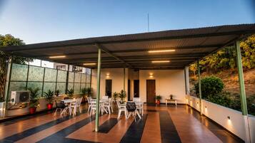 Un área de patio techada al aire libre con varias mesas y sillas, plantas en macetas y un equipo de aire acondicionado grande.