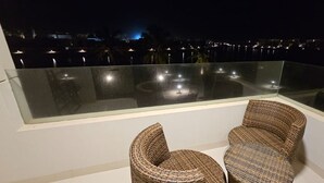 Een balkon met twee rieten stoelen en een glazen balustrade, met uitzicht op een stadsbeeld bij nacht.