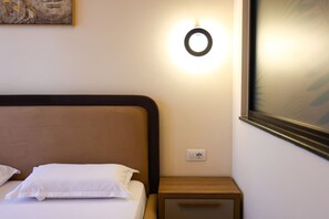 A neatly made bed with white linens and pillows, a wooden bedside table with a drawer, a wall-mounted light fixture, and a framed artwork above the bed. - Grandstay Urban Nine (Durrës)