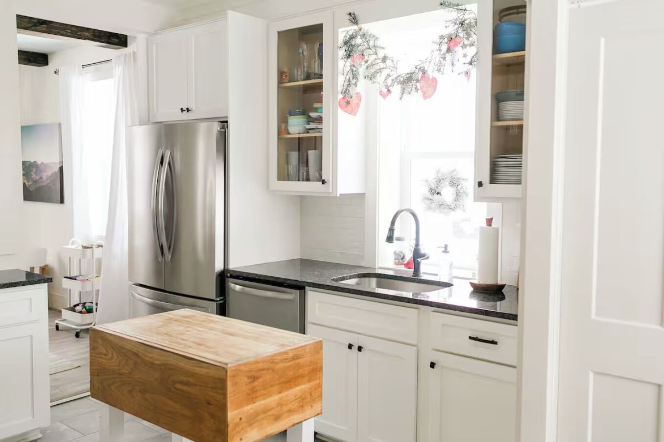A modern kitchen with white cabinetry, a stainless steel refrigerator, a wooden island, and a window with holiday decorations.