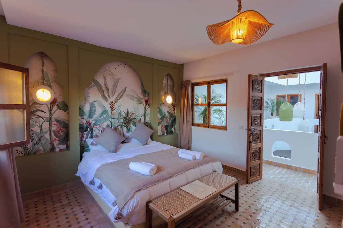 A bedroom with a bed, a bench, a wall with a tropical mural, and a view of a courtyard through an open door.