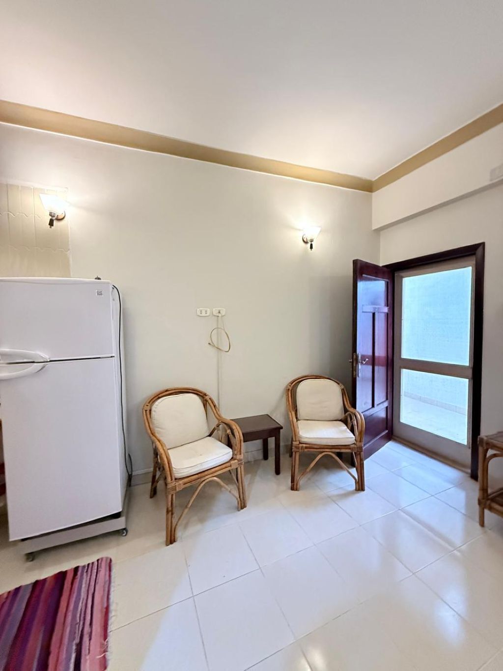 A compact living space with a white refrigerator, two wicker chairs, a small table, and a sliding glass door leading to a balcony.