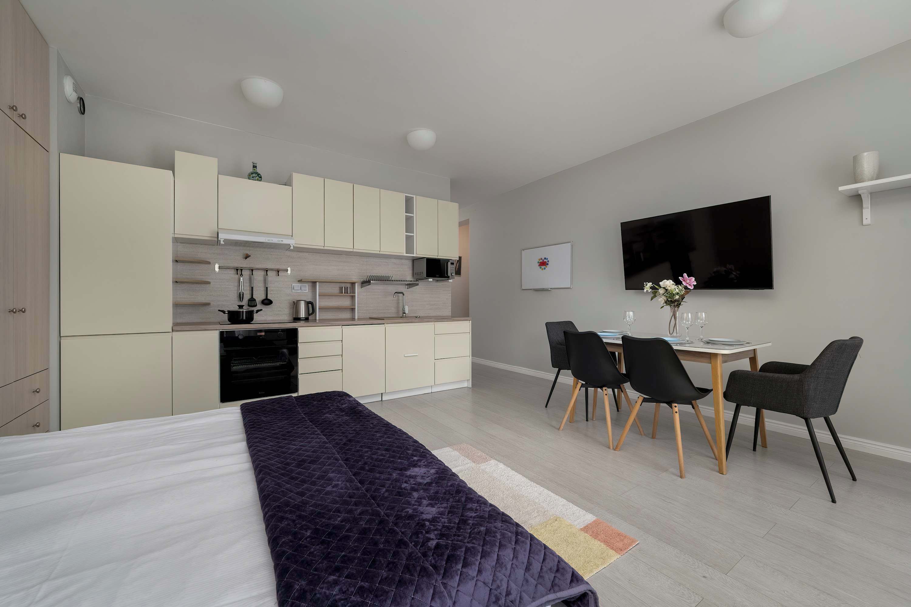 A modern hotel room with a bed, a dining area with a table and chairs, a kitchenette, and a flat-screen TV mounted on the wall.