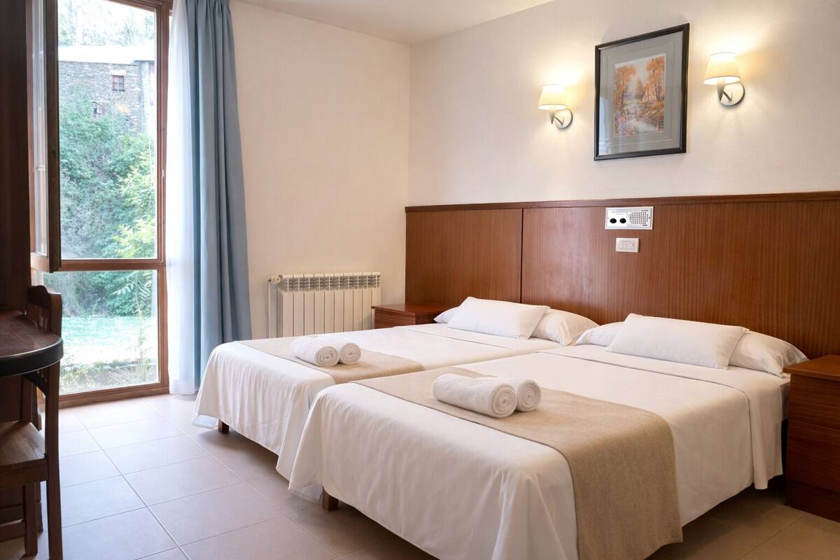 Ein Hotelzimmer mit zwei Betten, einem Holzkopfteil, einem Heizkörper, einem Schreibtisch und einem Fenster mit Vorhängen.