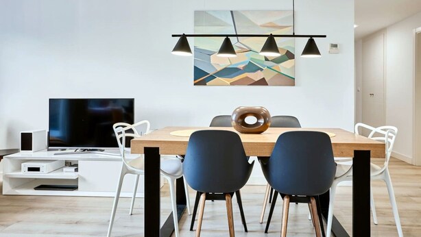 Een moderne eethoek met een houten tafel, zwart-witte stoelen, een flatscreen televisie en een wanddecoratie met geometrische vormen.