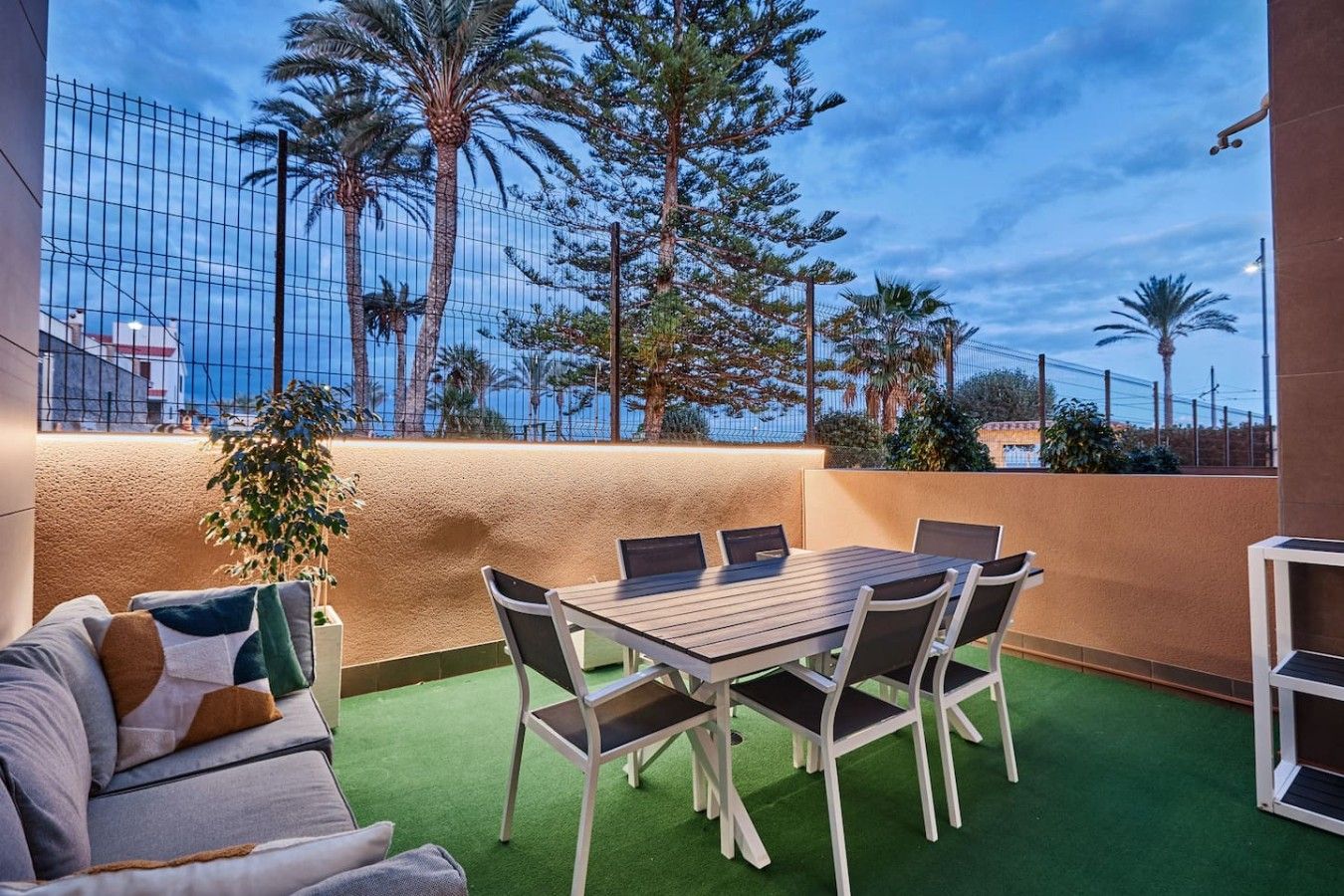 An outdoor patio with a table and chairs, surrounded by a low wall and a view of palm trees and a building.