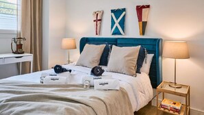 Een netjes opgemaakt bed met een blauw hoofdbord, twee nachtlampjes en een klein tafeltje met een boek.