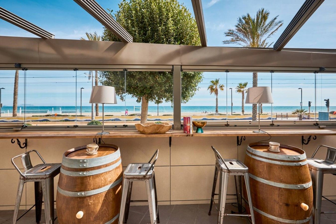 Un espace bar agrémenté de barils en bois, de tabourets en métal et offrant une vue sur la plage.
