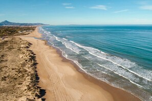 A coastal landscape with a sandy beach, ocean waves, and distant mountains. - Las Dunas 215 (Oliva)