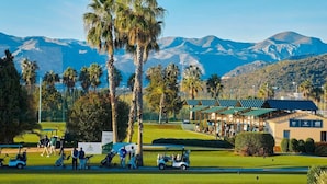 Een golfbaan met palmbomen, een clubhuis en bergen op de achtergrond.