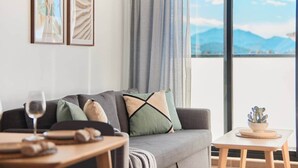 Een moderne woonkamer met een grijze bank, een houten tafel en uitzicht op bergen door een groot raam.