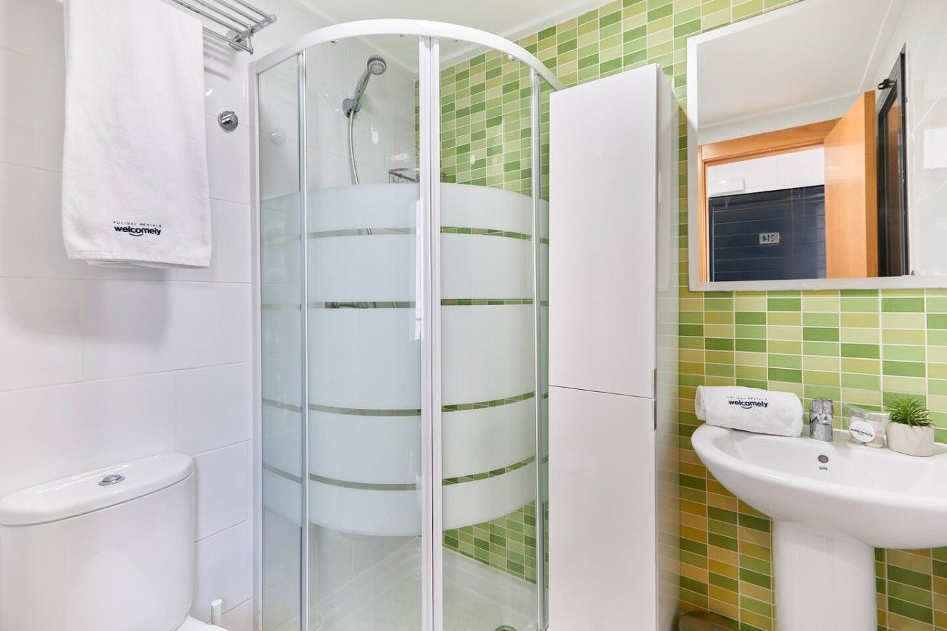 Un bany amb dutxa trencada per vidre, un vàter, un lavabo amb mirall i un penjador de tovalloles.