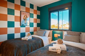 A hotel room with two beds, checkered walls, a window with a view, and a poster on the wall. - Las Dunas 515 (Oliva)