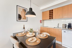 A modern kitchen with wooden cabinets, a built-in dishwasher, and a dining area with a dark table set with placemats and a potted plant. - Las Dunas 214 (Oliva)