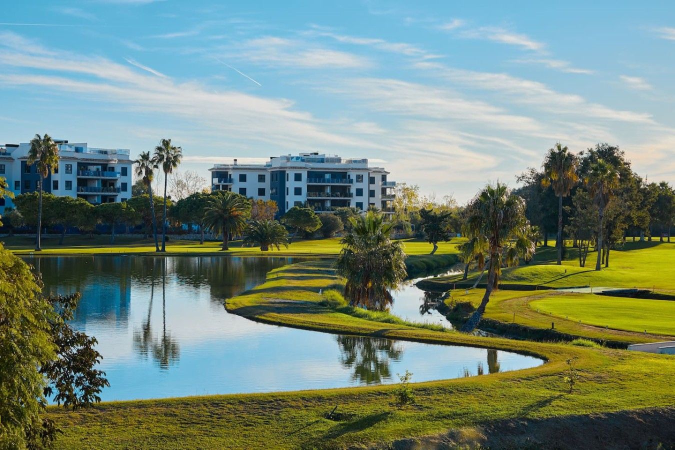 Campo de golf con un estanque, palmeras y edificios modernos al fondo.