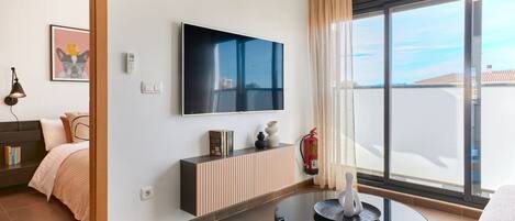 Habitació d'hotel moderna amb un televisor gran, un sofà, una taula petita i vista a la ciutat.
