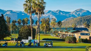 Ein Golfplatz mit Palmen, einem Clubhaus und Bergen im Hintergrund.