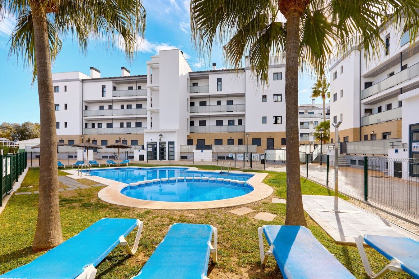 A pool area with blue lounge chairs, surrounded by a building with multiple balconies and a clear sky.